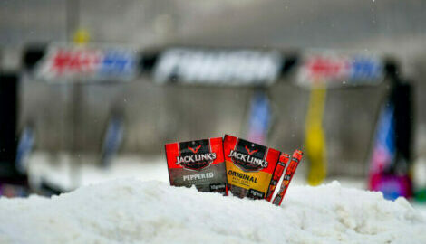 Three assorted bags of jerky in the snow with an out-of-focus snowmobile and finish line in the background