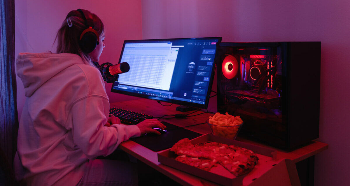 Young woman gamer sitting at a desktop computer and eating snacks in room with neon LED lights.